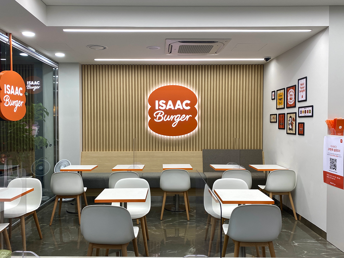 ISAAC Burger interior with an orange sign in the middle of the wall and white tables and chairs in two rows