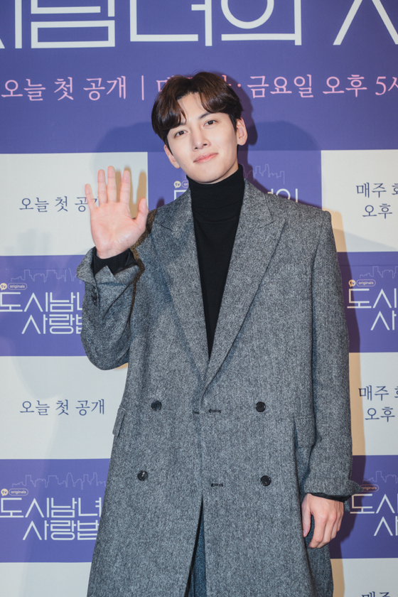 グレーのコートを着た男性がカメラに向かって手を挙げている様子。背景には韓国語で書かれた看板が見える。