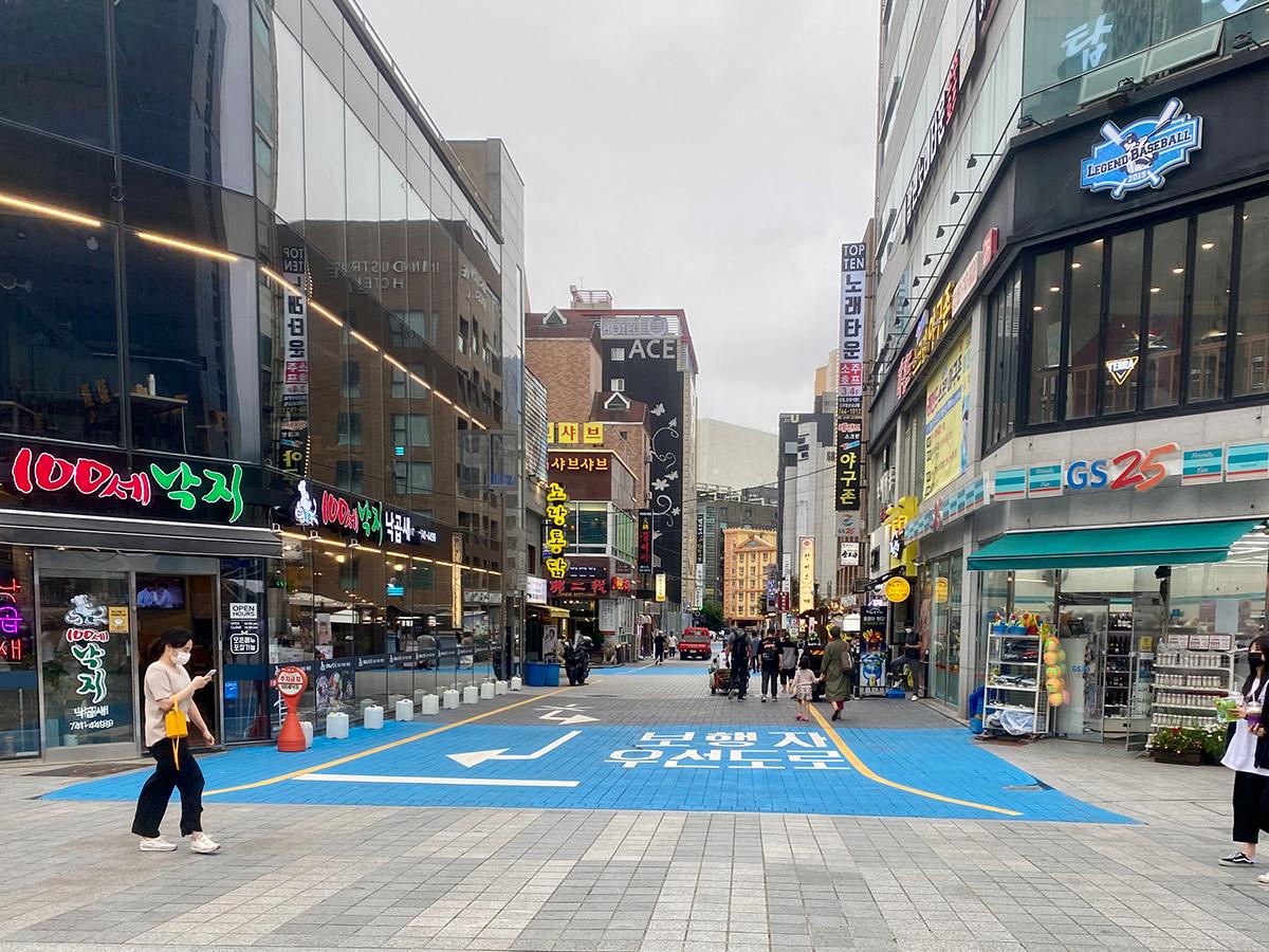 Persimpangan dekat GS25 di Busan, menunjukkan beberapa orang berjalan di jalan dengan toko-toko dan kafe berjejer di sepanjang jalan.