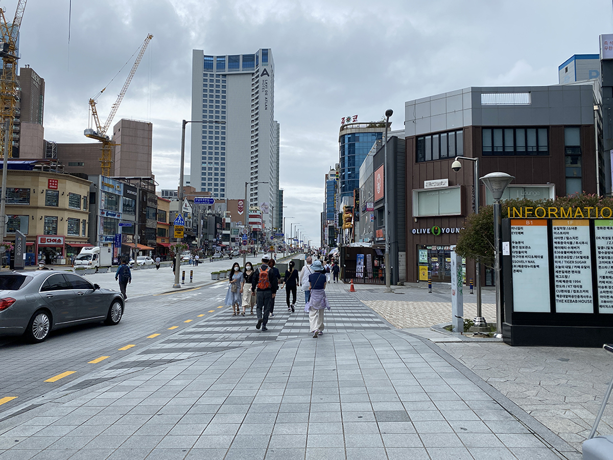 Jalan utama menuju Stasiun Haeundae di Busan, dengan pejalan kaki dan mobil di jalan, tampak beberapa gedung tinggi berjejer.