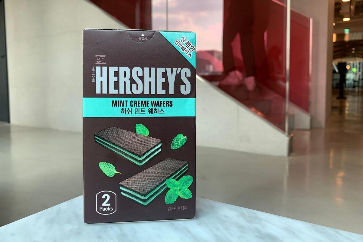 ภาพกล่อง HERSHEY’S Mint Creme Wafers ที่วางอยู่บนโต๊ะ ข้างกล่องมีรูปลูกอมมิ้นท์และเวเฟอร์โชว์ให้เห็นถึงรูปลักษณ์ของผลิตภัณฑ์