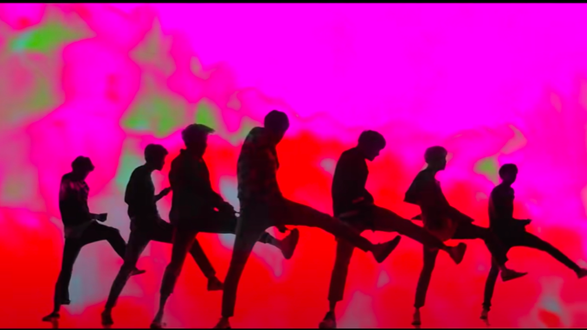 一群韓國偶像站在舞臺上，身著時尚的演出服，迎聚舞臺燈光的照耀。