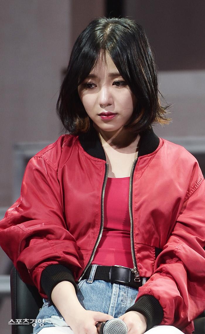 赤いジャケットを着た女性が座っている写真。彼女は青いジーンズを履いている。場面は室内で、控えめなライティングの下、彼女の衣服の質感が鮮明に見える。