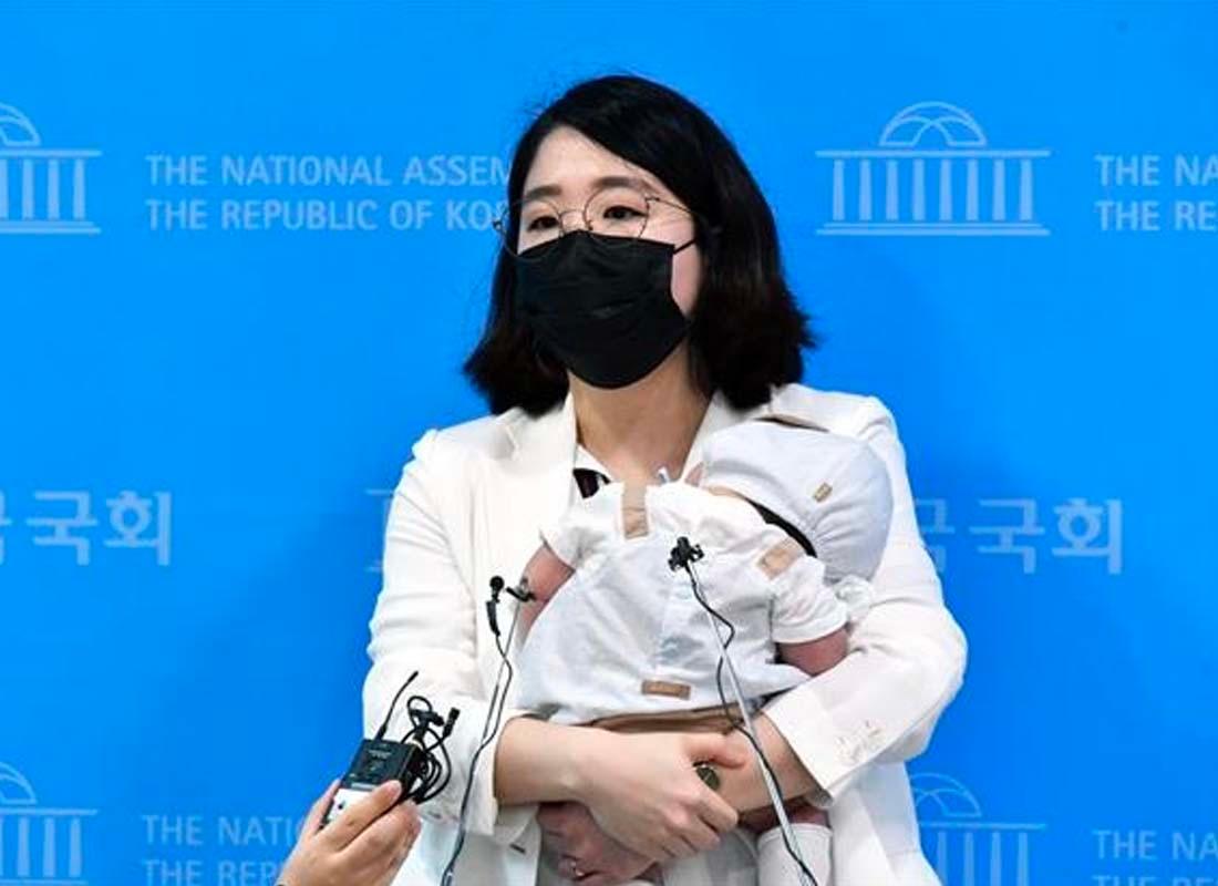 นักการเมืองหญิงกำลังแถลงข่าวพร้อมกับลูกในอ้อมกอด เธอสวมชุดสูทสีขาวและยืนต่อหน้าไมโครโฟนในรัฐสภาเกาหลี ภาพสะท้อนการส่งเสริมสิทธิสตรีในการมีส่วนร่วมทางการเมือง