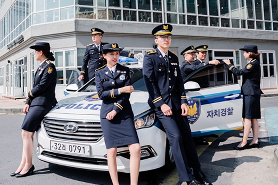 穿着韩国警察制服的人站在警车旁，背景是现代建筑，展示韩国警察现代化形象。