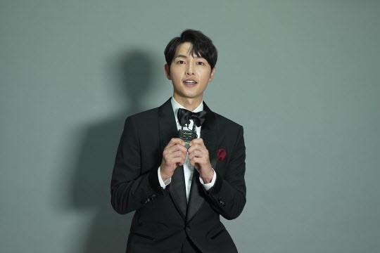 俳優が黒いタキシードを着て蝶ネクタイを結び、灰色の背景の前でトロフィーを持っている。