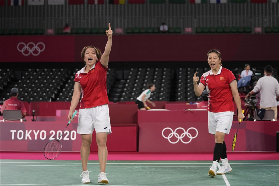 兩名中國羽毛球選手於2020東京奧運羽毛球賽場上慶祝，表現出勝利的喜悦與團隊默契。背景可見賽事場地的標誌性紅色設計。