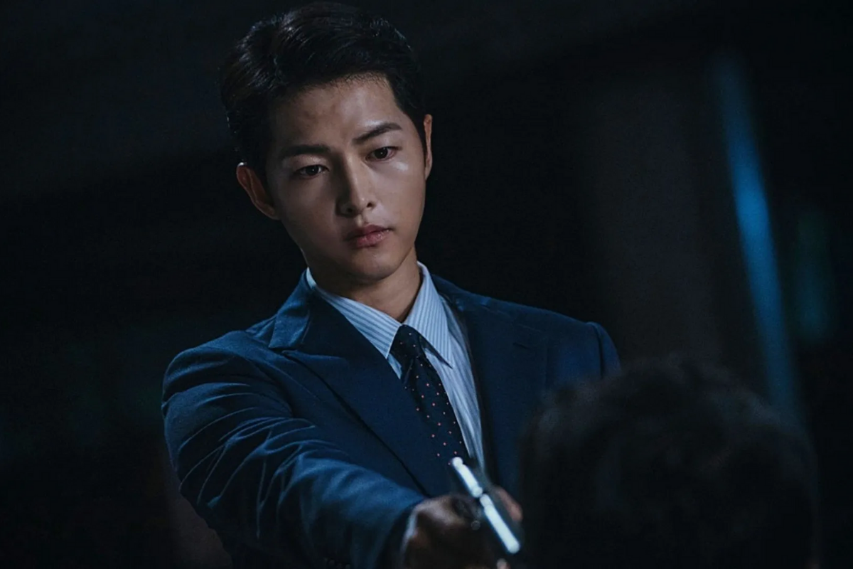 Hình ảnh từ phim Vincenzo, với nhân vật chính Song Joong Ki mặc vest xanh, đang cầm súng.