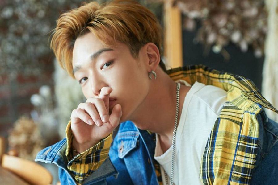 iKON组合成员Bobby，穿着黄色格子衬衫和牛仔夹克，坐在木制背景前。