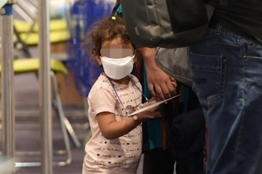 一个身穿口罩的小女孩紧握着亲人的手，在机场等待。