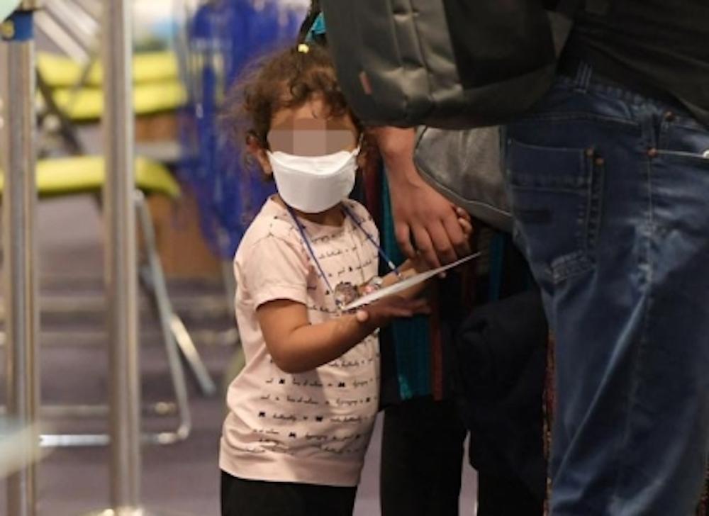 Afghan refugee child at Incheon Airport holding a stuffed animal, accompanied by security guards in quarantine suits.