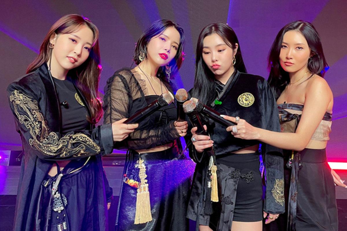 韓國女團 Mamamoo 穿著韓服風格的禮服，手中握著麥克風，背景是舞台燈光，展現出獨特的魅力。