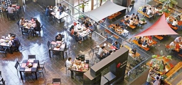 ビジネスマンたちがオフィスビルの軽食コートで昼食をとる様子。テーブルは安全距離を保って配置されており、食事を楽しんでいる様子が散見されます。