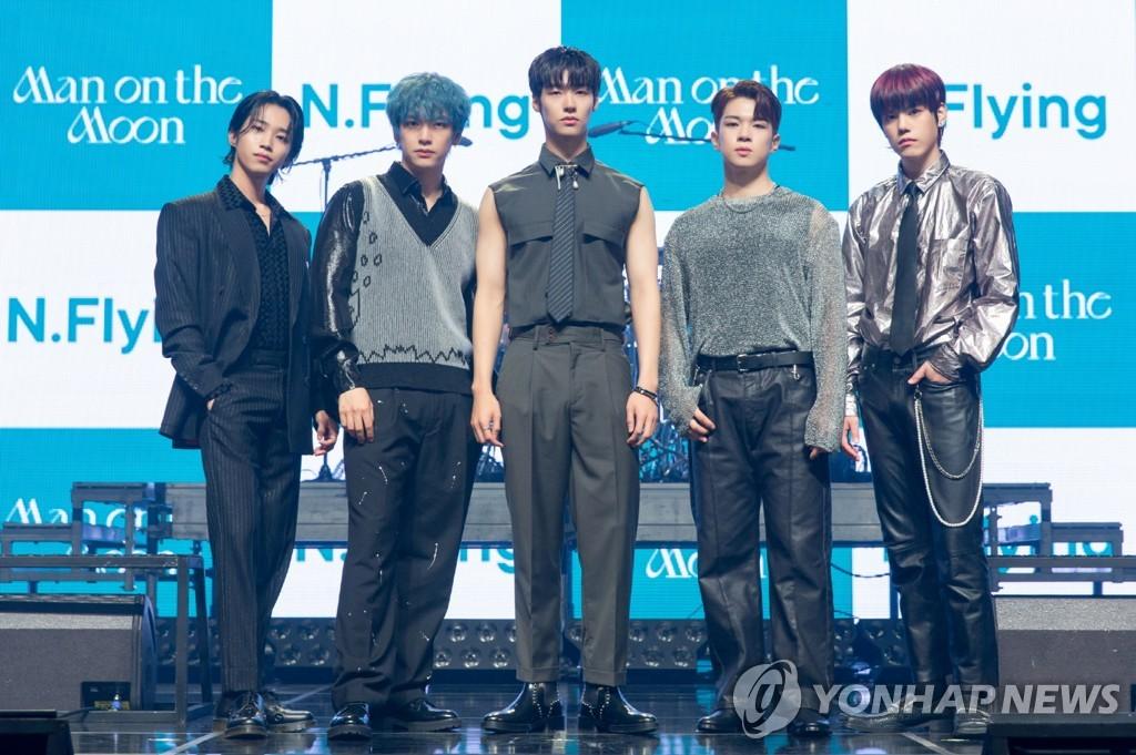 N.Flyingのメンバーがステージで公式の場に立っているシーン。新型コロナウイルス感染症に関するニュースの一部。