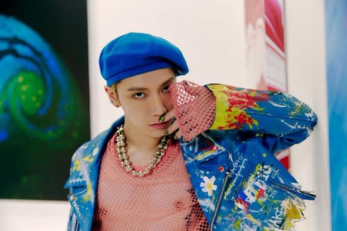 ศิลปินชื่อ Ten จากวง NCT สวมหมวกเบเร่ต์สีน้ำเงินและเสื้อแฟชั่นแบบเปิดด้านในพร้อมสร้อยคอโซ่ในฉากหลังที่มีหน้างานศิลปะที่มีสีสันสดใสในแกลเลอรี.