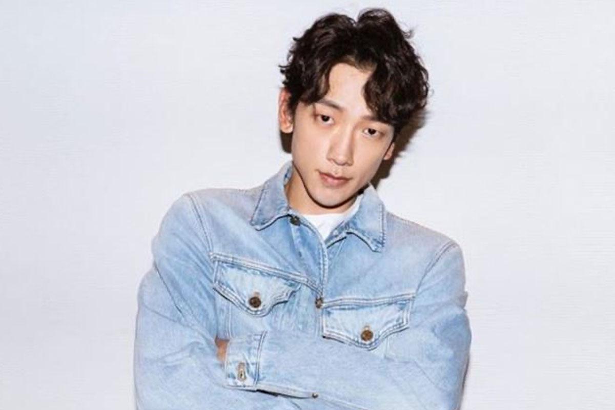 ศิลปินชื่อ Rain สวมเสื้อแจ็คเก็ตยีนส์สีฟ้าพร้อมแขนพับขึ้นครึ่งหนึ่งยืนถ่ายรูปในสตูดิโอที่มีฉากหลังสีขาวสะอาด.