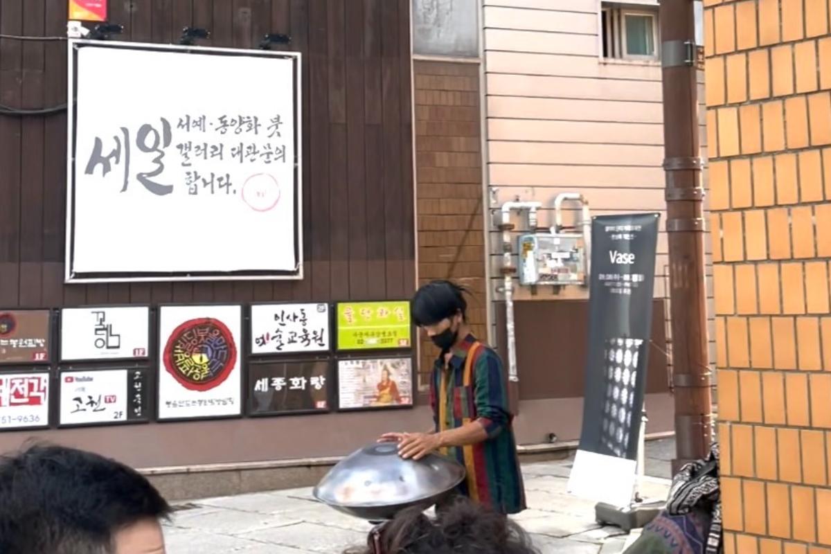 Artiste de rue jouant de la musique à Insadong, Séoul, devant un mur avec des affiches et des signes culturels, créant une ambiance vivante et artistique.