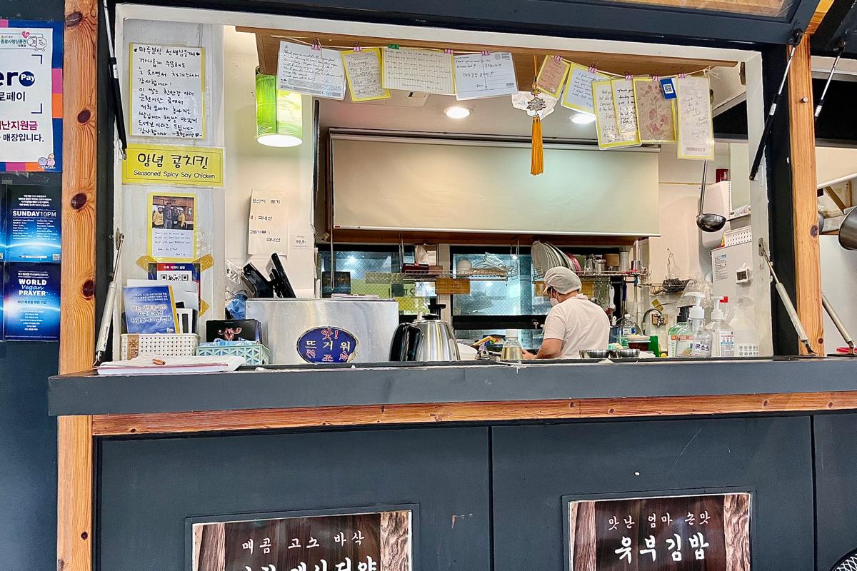 Employé du snack-bar Maru JaYeonSik Kimbap à Insadong, préparant les plats pour les clients dans une cuisine ouverte et accueillante.