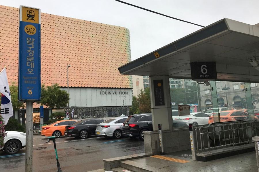 ภาพสถานีรถไฟใต้ดิน Apgujeong Rodeo ในกรุงโซลที่มีป้ายกำกับทางออก 6 เด่นชัด ใกล้กับถนนเส้นหลักที่มีรถยนต์จอดอยู่บางส่วนและมีความคึกคัก
