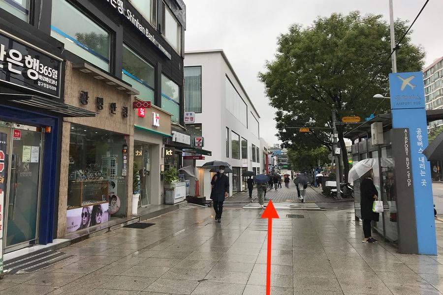 ภาพถนนที่มีคนเดินไปมาใกล้กับทางออกของสถานีรถไฟ Apgujeong Rodeo ในกรุงโซล ฝนตกทำให้บริเวณพื้นเปียก ถนนเต็มไปด้วยร้านค้าที่มีป้ายโฆษณา