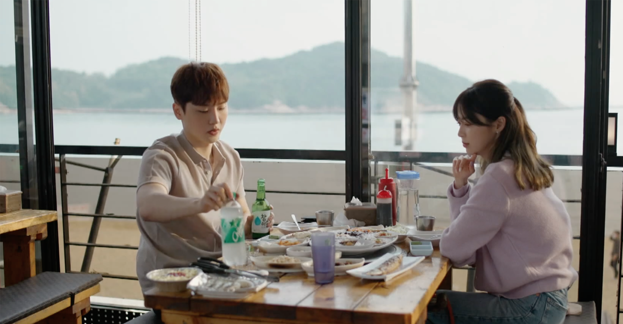 兩人在海邊餐廳用餐，桌上的飲料和海鮮佳餚豐富，背景是一片風景優雅的海灘。