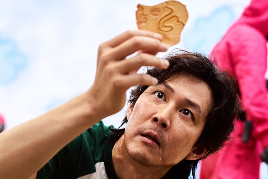 參賽者正拿著一塊形狀獨特的椪糖，周圍是穿粉紅色衣服的警衛，《魷魚遊戲》其中一幕，來自Netflix。