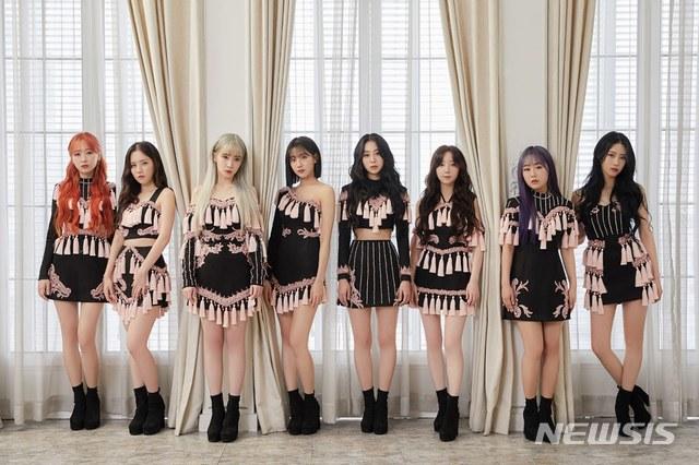 Lovelyzのメンバーがウリムエンターテインメント提供の衣装を着て並んで立っている。メンバーたちはほぼ同じデザインの黒とピンクの衣装を着用し、背景には大きなカーテン付きの窓がある。