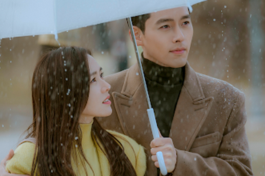 韓劇《愛的迫降》中的場景，畫面中兩位主角撐著白色傘，背景有雪花飄落，展現浪漫氛圍。