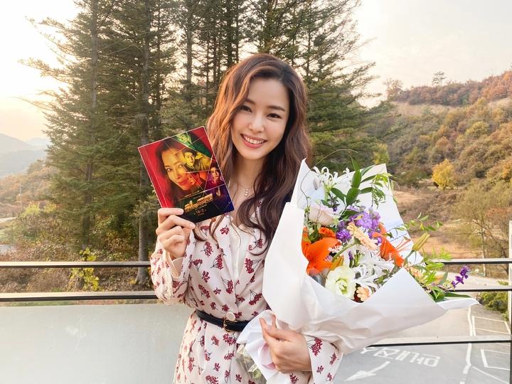 美しい風景の前で花束と小冊子を持つ女性。女性は、花柄の洋服を着ており、手には目立つ花束とカラフルなデザインの小冊子を持っています。背景には緑豊かな自然の景色が広がっています。