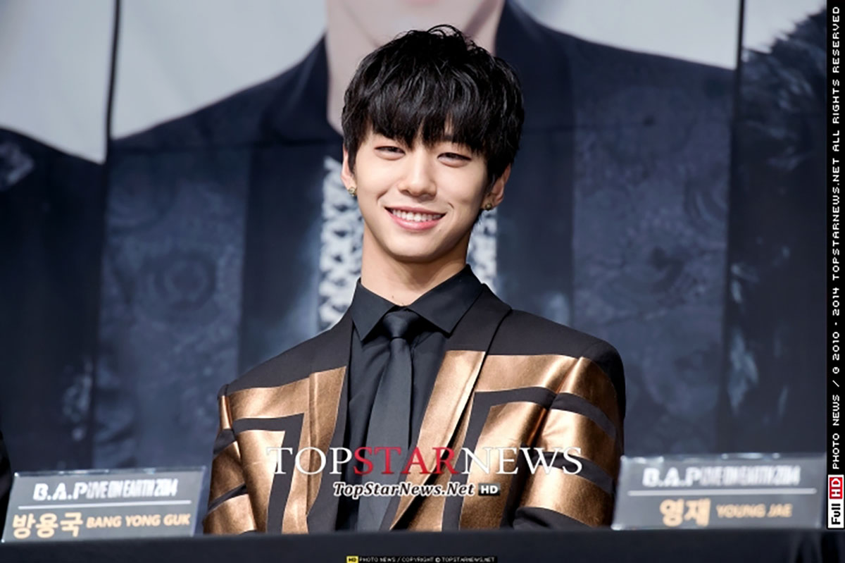 B.A.Pのバン·ヨングクがフォーマルなスーツを着て、記者会見で出演者やファンに向けて話している。彼はプロデューサーとしての活動も知られている。