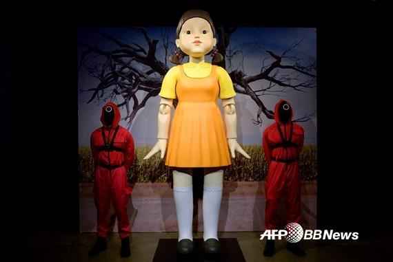 黄色の制服を着た巨大な人形と赤い衣装のフロントマン、握手する道具が展示された、韓国ドラマ『イカゲーム』のセット