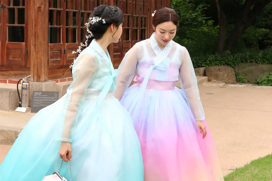 两名穿着浅蓝色和粉紫色韩服的女性站在传统韩国建筑前，其中一人手持一个包。