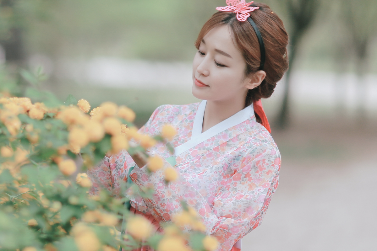 Một phụ nữ mặc hanbok màu hồng đứng gần một bụi hoa vàng, trang phục truyền thống Hàn Quốc mang lại vẻ đẹp dịu dàng và lãng mạn trong khung cảnh tự nhiên.