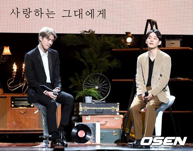 EXOのチェンとセフンが音感会のステージでマイクを手にして座っている。背景には韓国語で書かれた文字が飾られている。
