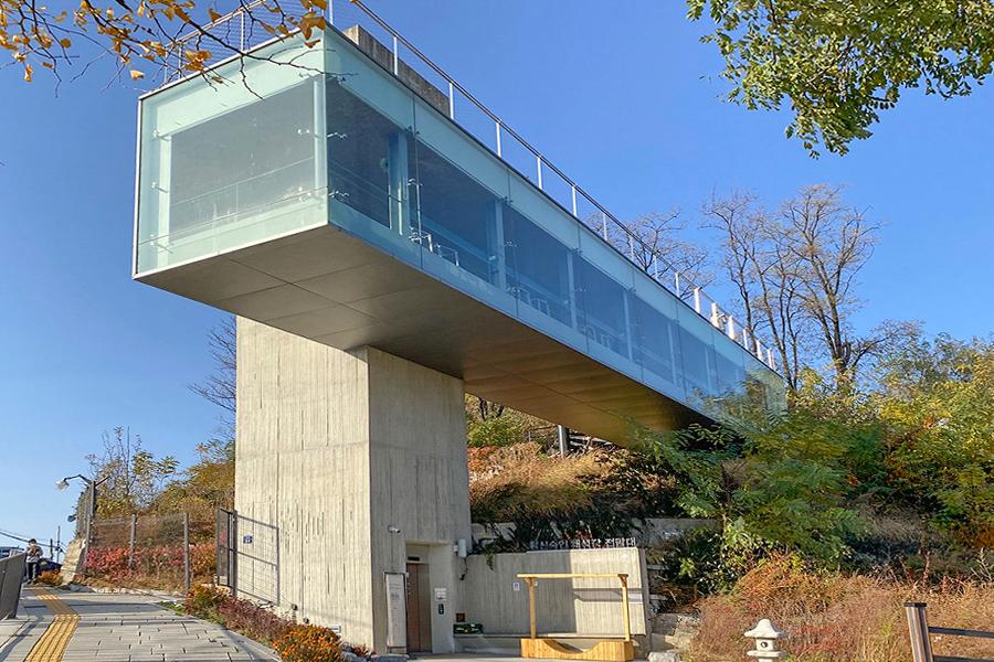 昌信洞崇仁采石场展望台的外观,透明玻璃设计的建筑物坐落在青翠的树木之间,映衬蓝天白云,吸引游客来到这个首尔著名的景点欣赏美景。