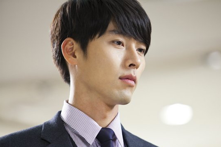 身穿西裝、搭配襯衫和領帶的男子站在室內，背景模糊。