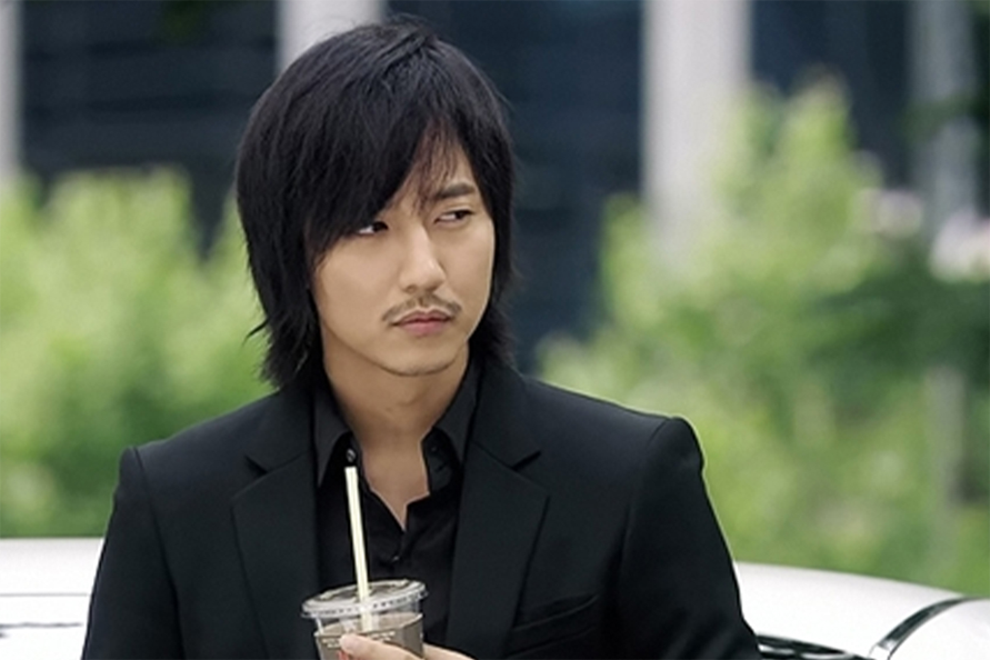 身穿黑色西裝的男子在戶外拿著飲料杯，背景是淺色的建築物。