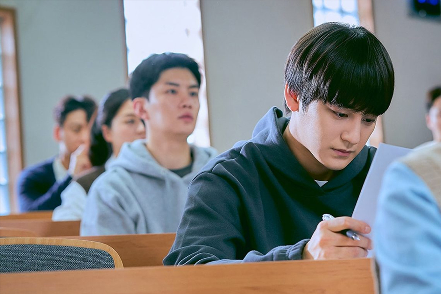 男性在閱讀文件，身穿深色連帽衫，背景模糊的教室場景。