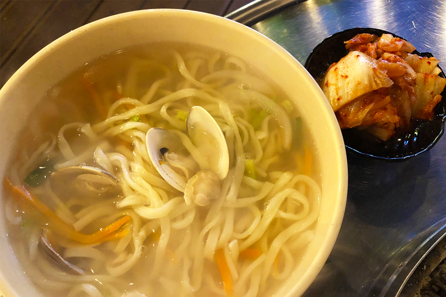 A steaming bowl of handmade noodle soup with seafood at Dalbit Shellfish Grill in Wolmido, Incheon, accompanied by side dishes.