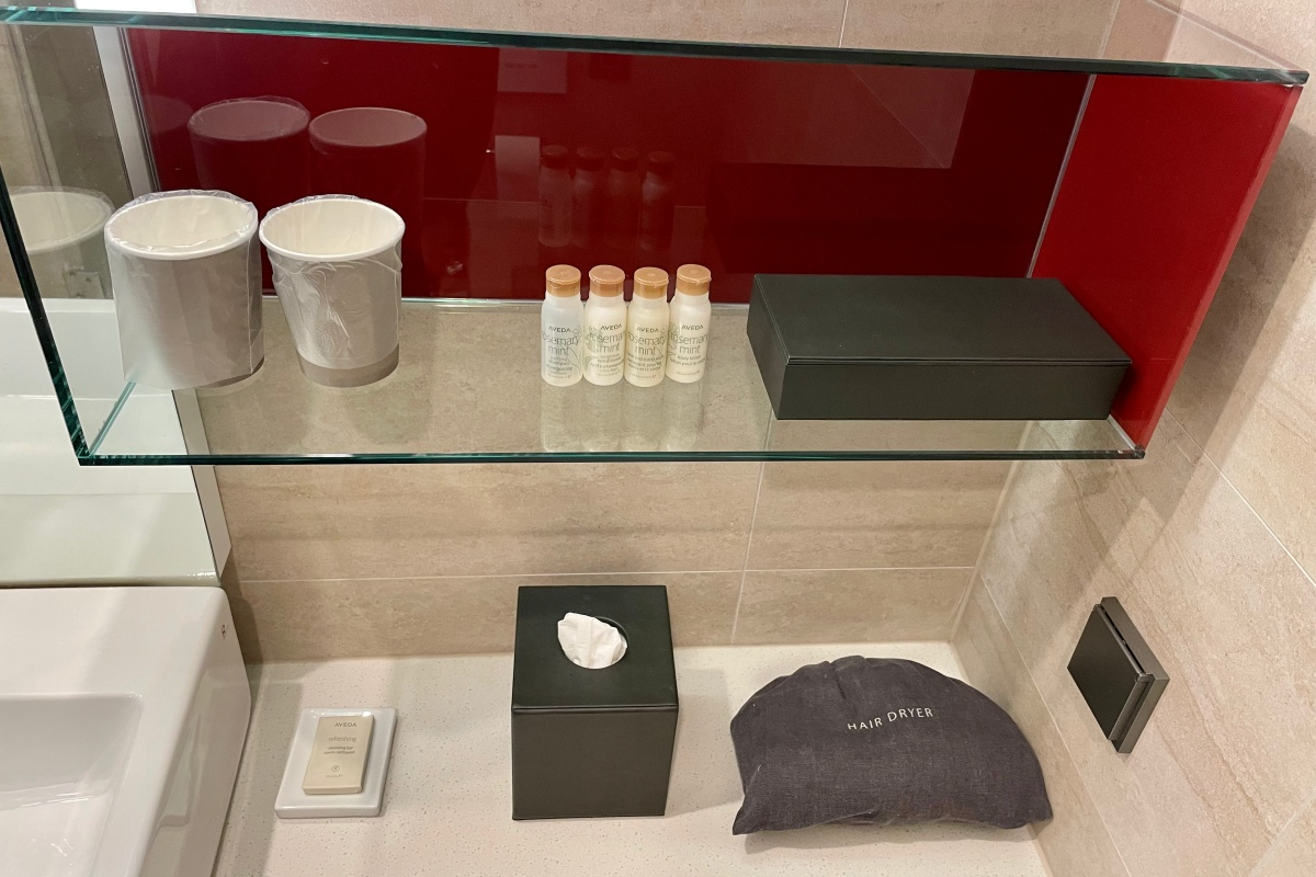A shelf holding two paper cups in plastic bags, some shampoos and lotions, a box, some tissue, and a blow dryer.