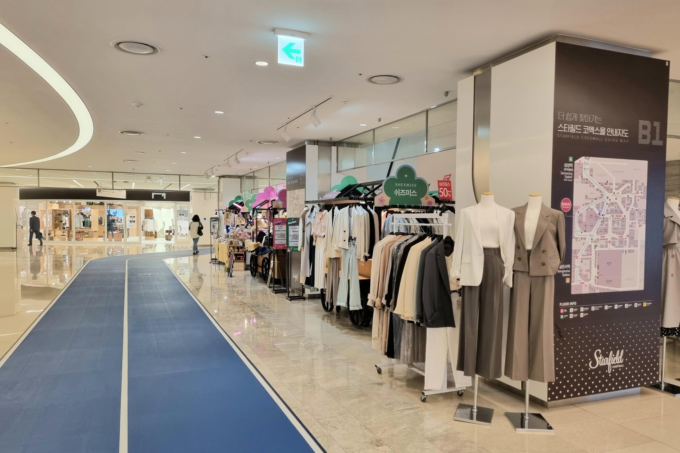 코엑스몰 내부의 쇼핑몰 레이아웃을 보여주는 사진입니다. 현대적인 실내 디자인과 밝은 조명이 공간을 한층 더 돋보이게 하며, 다양한 패션 의류 매장과 디스플레이된 쇼핑 아이템들이 눈길을 끈다.