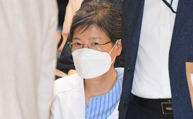 韩国前总统朴槿惠出庭，身穿白色上衣，身旁有工作人员陪同。