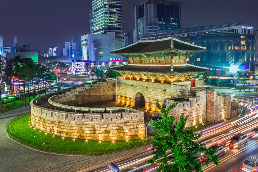 서울의 밤에 촬영한 동대문 전경. 화려한 조명이 켜진 동대문과 주변의 건축물들이 보입니다. 차들이 바쁘게 지나가는 모습이 인상적입니다.