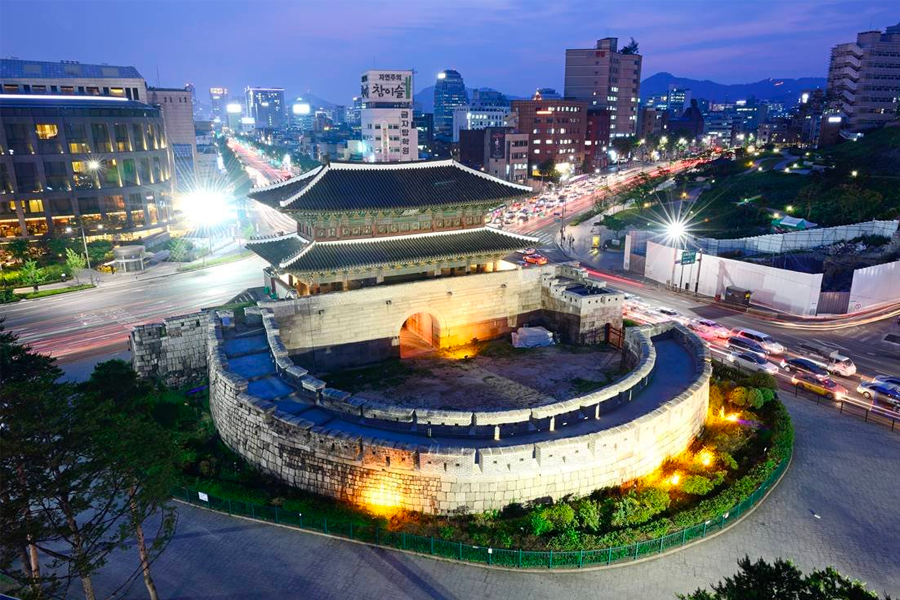 황혼 무렵 서울 동대문의 전경. 조명이 켜진 동대문 앞쪽으로 차량의 불빛이 거리의 선을 그리고 있습니다. 도시의 활기가 느껴지는 모습입니다.