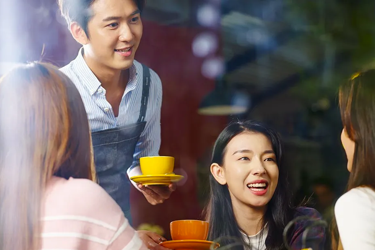 臺北的一家咖啡廳內有服務生端著一杯黃色飲品和盤子，幾位顧客正在聊天和享用咖啡的情景。