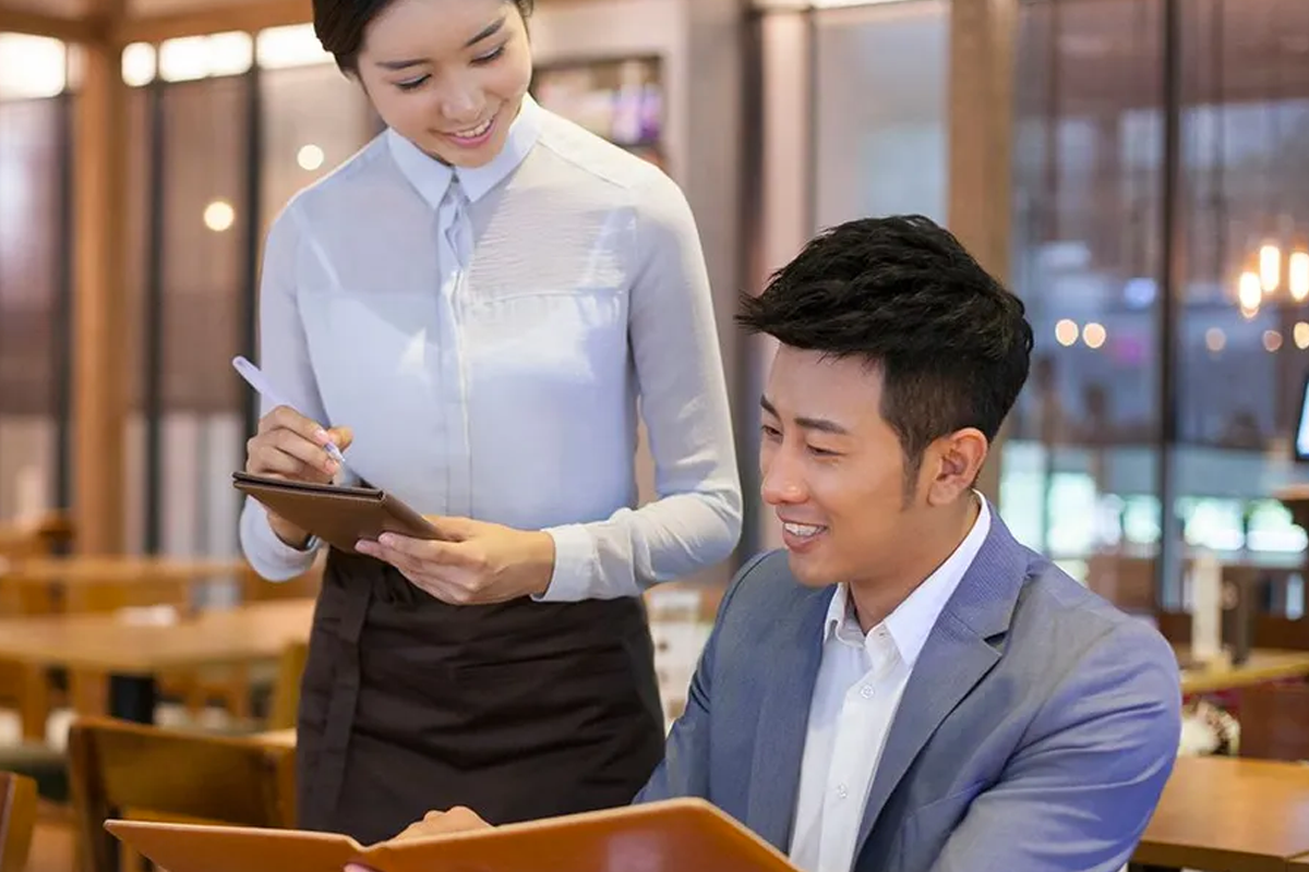 一家餐廳內服務生與顧客互動的畫面，顧客正查看菜單，服務生在一旁準備記錄點餐。