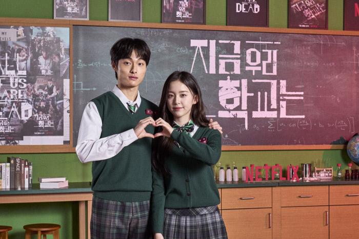Netflixシリーズ「今、私たちの学校は...」の学生役の俳優が黒板の前にカップルのようにポーズ。教室の一部が背景に見える。
