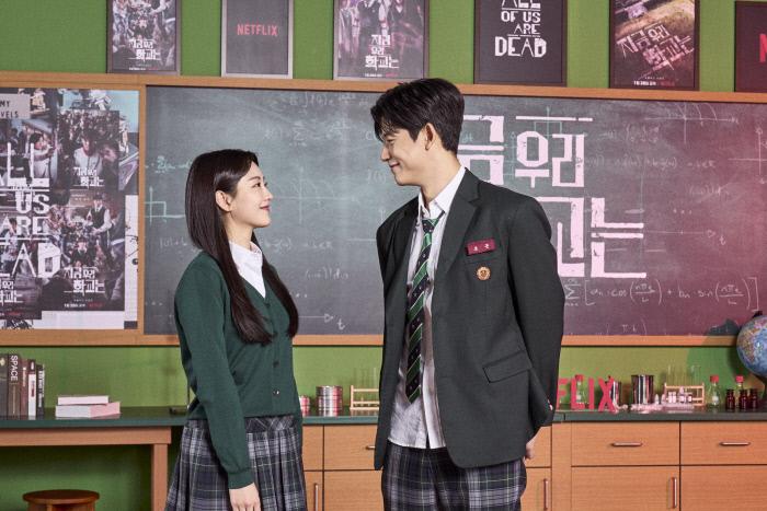 Netflixシリーズ「今、私たちの学校は...」の制服を着た出演陣が教室で撮影。ポスターが壁に並んでいる中で、真剣なまなざしを交わすシーン。