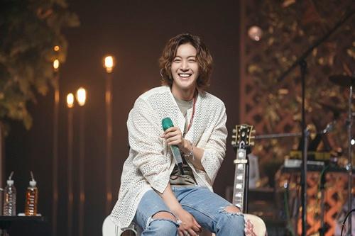 コンサートで観客にメッセージを伝えるキム・ヒョンジュン、ステージ上で落ち着いた雰囲気の室内に座りながらマイクを持ち、ギターが横に置かれている姿。