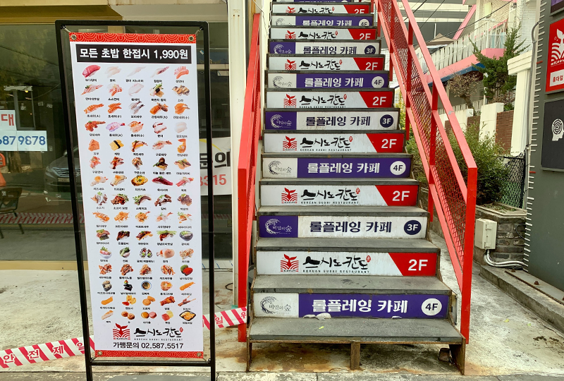 通往Sushi Nokando壽司店的樓梯，臺階上貼有店名廣告，旁邊有壽司菜單海報。